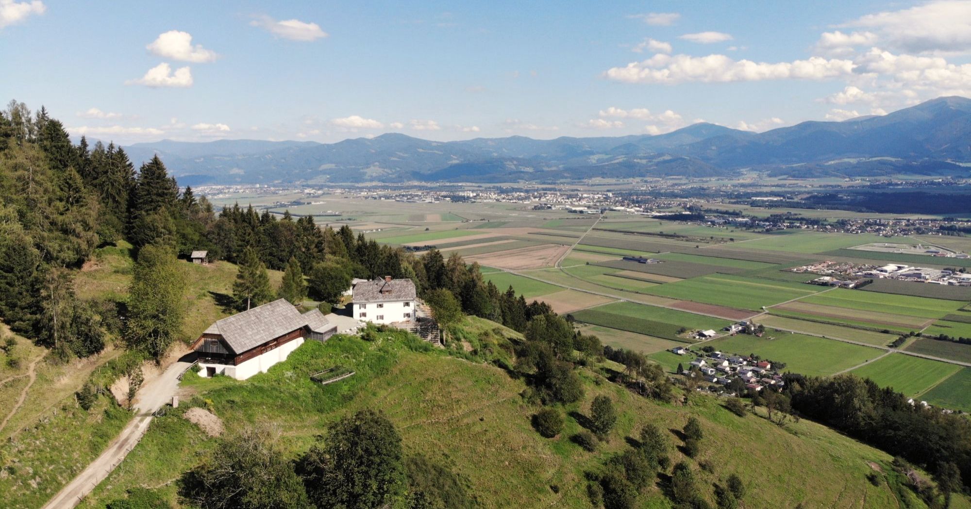 Luftbild: Stadl und Wohnhaus auf dem Hügel, darunter die Felder des Aichfelds, dahinter die Berge