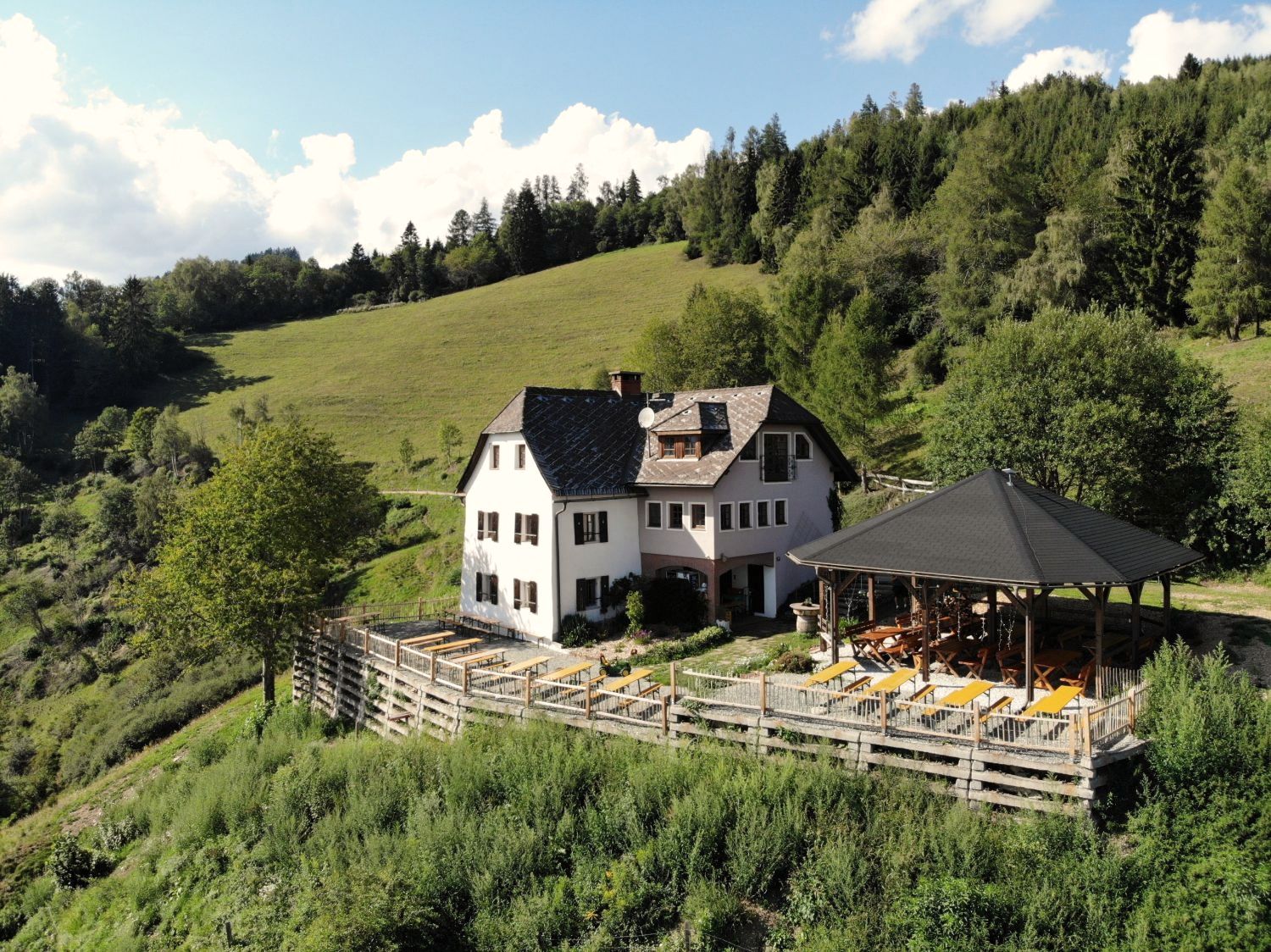 Luftbild: das weiße Wohnhaus mit Holzterrasse und Pavillon am Waldhang