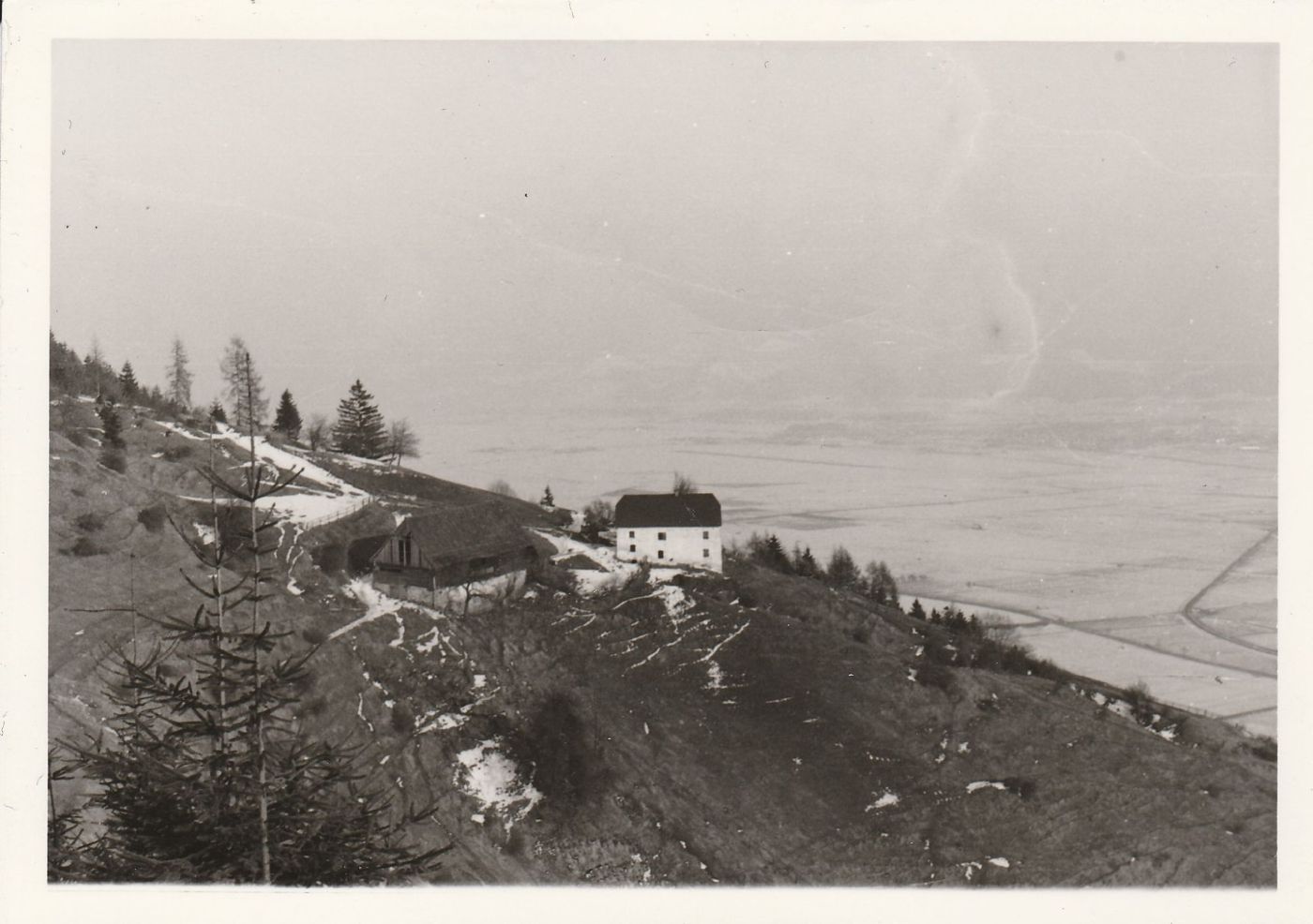 Altes Schwarzweißfoto: der Hof am verschneiten Hang, unten das Tal