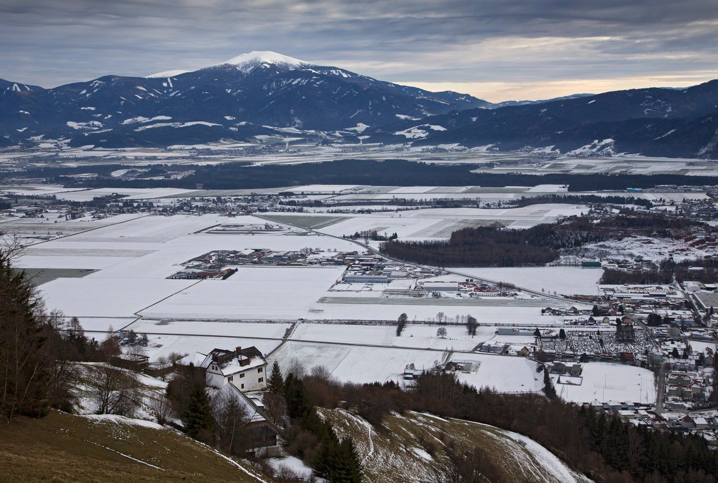 Winterpanorama: das Wohnhaus am Waldrand, darunter das verschneite Aichfeld und ein schneebedeckter Berg